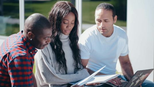 Three colleagues collaborating outdoors, discussing documents and using a laptop.